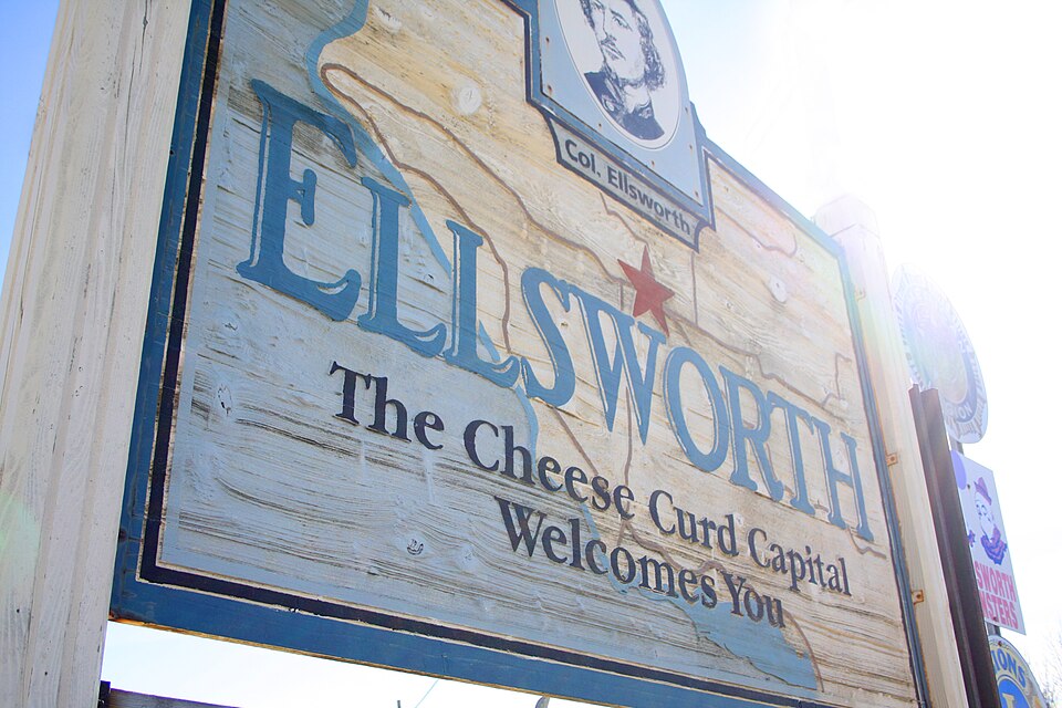 Wooden welcome sign reading "Ellsworth, The Cheese Curd Capital Welcomes You," with community organization emblems on either side., Pierce County, Wisconsin
